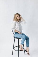 Beautiful blonde girl in white shirt and jeans sitting on a chair on a white background in the studio.