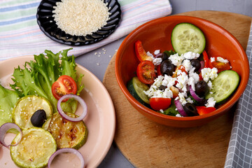 beige container with grilled zucchini on lettuce leaves and greek salad on a gray background on a blue napkin with a fork near pepper and onion. Horizontal photo