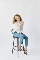 Beautiful blonde girl in white shirt and jeans sitting on a chair on a white background in the studio.