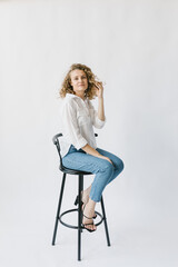 Fototapeta premium Beautiful blonde girl in white shirt and jeans sitting on a chair on a white background in the studio.