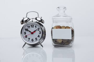 Coins in a glass bank with space for text and alarm clock in a white room. Time is money.