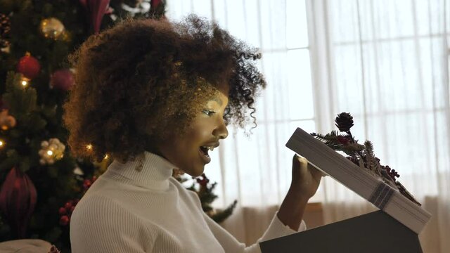 Nice young lady with dark skin and kinky hair opens present box and smiles widely against Christmas tree closeup slow motion