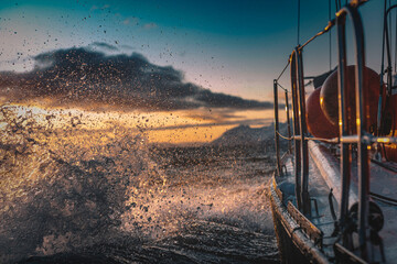 Sail boat sailing through rough sea water with waves splashes, side view with snowy mountains on...