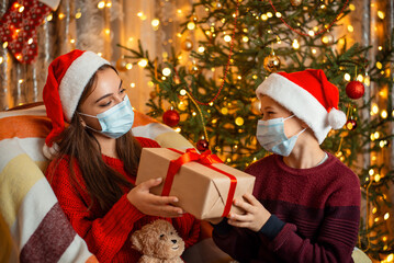 Fototapeta na wymiar Girl sharing box gift with her younger brother, both in medical masks. Holidays with family in isolation, new year tree and lights with decorations on the background.