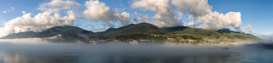 Fototapeta na wymiar Panoramic shot of mountains covered with clouds and fog in Gastineau Channel, Juneau, Alaska. Golden hour. Blue cloudy sky as a background and boat sailing in the fog in the foreground