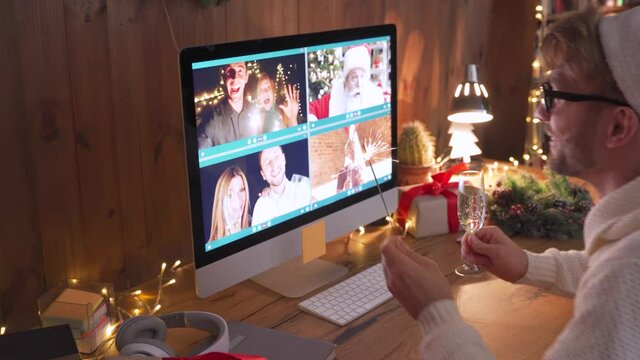 Young man wearing Christmas hat drinking champagne holding sparkler talking to friends on virtual video zoom call celebrate Happy New Year party in distance online conference chat on computer at home.