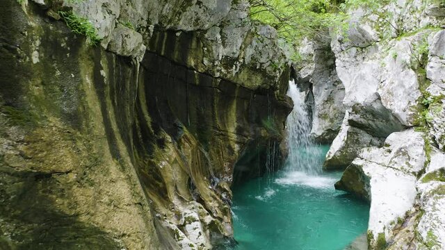 Mesmerizing view of Turquoise/Emerald Water of Soca River Flowing through the narrow Gorge between huge Rocky Mountains With Waterfalls overflowing Soca River and Forest in Background.
