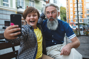 Happy grandfather and young grandson laughing, taking selfies on samrt phone outdoors in the city