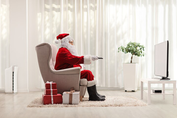 Santa claus in armchair with a remote control in front of tv at home with presents on the floor