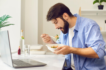 Young man sitting at workplace and eating healthy balanced delivered meal from container