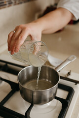 A hand of a woman who is pouring fresh lemon juice from the glass to the saucepan on the gas stove