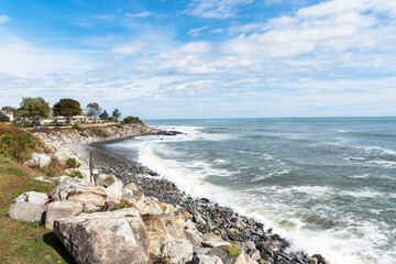 Small pebble beach along the coast of New Hampshire on a sunny autumn day. A clifftop coastal road lined with houses is visible in background