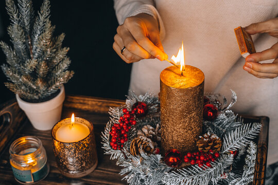 woman hands lightning big gold candle with matches in Christmas wreath. Xmas interior decoration on rustic wooden background. hygge, ornaments, candle burning in lantern on window sill at home.