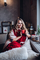 Pretty blonde girl in red wrapped in plaid enjoying Christmas morning with mug of coffee by window.