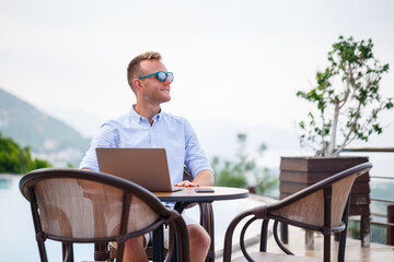 Successful handsome male businessman in sunglasses works at a laptop sitting near the pool. Remote work. Freelancer