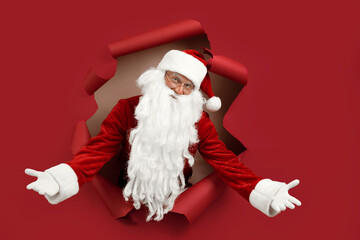 Santa man spread his arms in different directions and look emotionally through a paper hole. Bearded man in santa hat looking through hole on red paper.