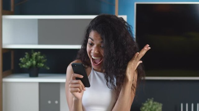 Happy african american woman looking in smartphone feel surprised, saying WOW and makes Yes gesture. Celebrating success victory. Excited black female winner hold mobile phone amazed with good news.