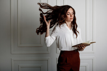 A beautiful young girl reading a book and throwing long wavy hair up. Model in a white blouse and red trousers in office style clothes. Space for text.