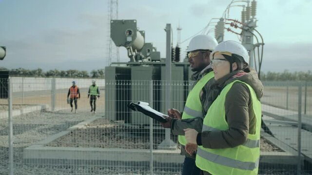 Engineers man and woman in warm clothes hold black folder and discuss work walking against power electric substation closeup