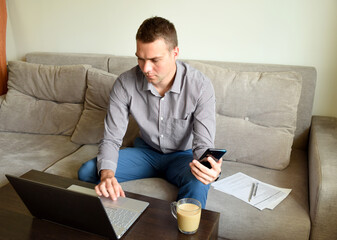 Young tired business man in shirt and trousers sitting on the couch and working with pensive serious face behind laptop after work in office. he holding phone.concept of working from home 