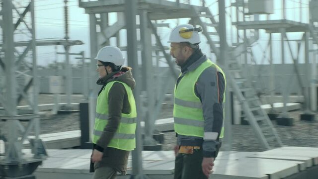 Electrical engineers in reflective vests climb on ladder to industrial power station transformer cabinet ground and discuss work