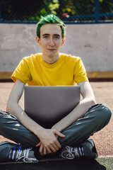 Freelancer, it professional in free space, remote working at online platform for programmers. Smiling young male freelancer with green hairstyle working on laptop in outdoor street.