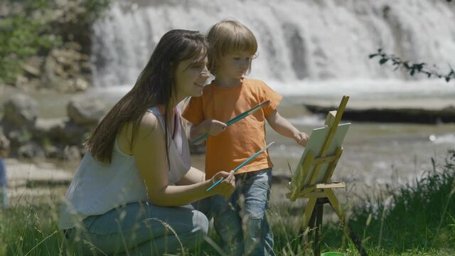 Portrit of mother and little child paint together on easel in nature, waterfall, creative summer holiday