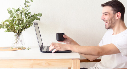 Retrato de un hombre adulto y apuesto caucásico sentado frente a su laptop y bebiendo una taza de...