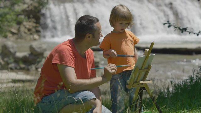 Father and little son painting together near waterfall flowing, inspirational nature, free creativity, summer holiday