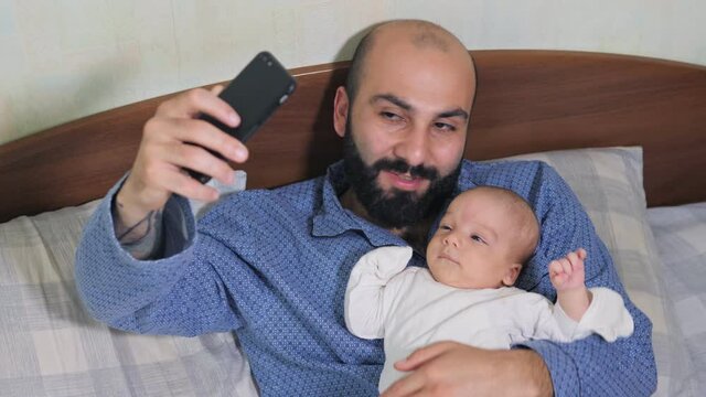 Close up of father and his newborn baby making video call to mother or relatives in a bed. Concept of technology, new generation, family