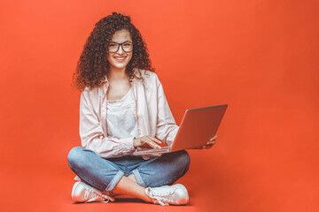 Happy young curly beautiful woman sitting on the floor with crossed legs and using laptop on red...