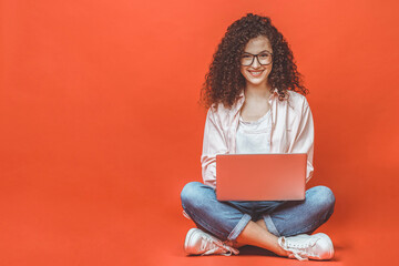 Happy young curly beautiful woman sitting on the floor with crossed legs and using laptop on red...
