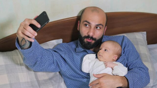 Close up of father and his newborn baby making video call to mother or relatives in a bed. Concept of technology, new generation, family