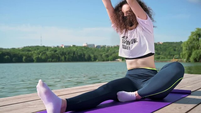 Young curly woman practicing yoga near the lake while wearing black protective mask. Sunny day. Meditating in lotus position on the yoga mat. Corona Virus idea