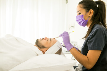 Obraz na płótnie Canvas Limpieza facial de un hombre calvo en un spa, con fondo blanco y guantes morados y mujer dermatologa con cubre bocas