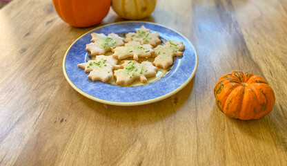 Above view of a plate of sugar cookies in the shape of maple leaves with green sprinkles on them. Cookies are arranged on a dark blue plate on a brown table with an orange sugar pie pumpkin nearby.