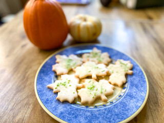 Above view of a plate of sugar cookies in the shape of maple leaves with green sprinkles on them. Cookies are arranged on a dark blue plate on a brown table with an orange sugar pie pumpkin nearby.