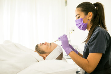 Obraz premium Limpieza facial de un hombre calvo en un spa, con fondo blanco y guantes morados y mujer dermatologa con cubre bocas