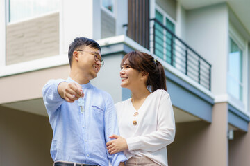 Fototapeta premium Portrait of Asian young couple standing and hugging together and holding house key looking happy in front of their new house to start new life. Family, age, home, real estate and people concept..