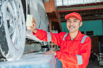 Male car cleaner wears red uniform and smiling hat while washing the bottom of the car in the car salon