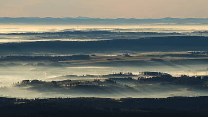 Sunrise light rays casting long shadows in the morning mist rolling over hilly landscape with wind turbines peaking from the clouds int he background.