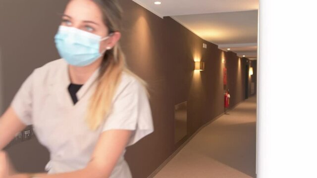 Maid with face mask and the floor trolley walks through a corridor in the hotel. Covid-19 tourism and hospitality footage.