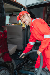 Asian male car cleaner wears red smiling uniform while cleaning car floor with vacuum cleaner in car salon