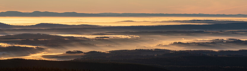 Landscape panorama with light rays from rising sun peaking through morning mist during inversion.