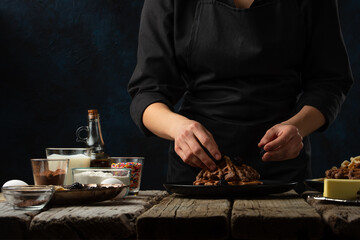 The chef in black apron adds blackberry on waffles. Backstage of serving waffle dessert for breakfast on rustic wooden table with ingredients on dark background. Frozen motion. Traditional recipe.