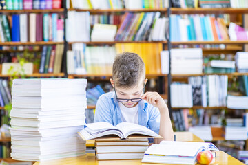 A boy with glasses sits at a table in a library with a stack of books and reads