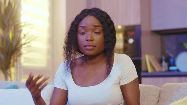 Serious beautiful african american young woman look at camera waving his arms twists his finger on his head sitting at home on sofa. Quarantine time. Stay at home. Close up. Slow motion