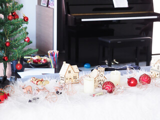 Christmas decorations of wooden houses, balls, candles, lights on white wool, with a piano backdropdr..