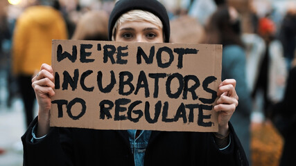 Womens march. Young woman with face mask holding banner sign - We are not incubators to regulate. protest against strict abortion laws. High quality photo - obrazy, fototapety, plakaty