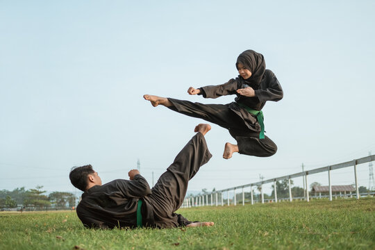An Asian male fighter in a pencak silat uniform with a kicking pose from the floor and a hooded female fighter in a flying kick in an outdoor background.
